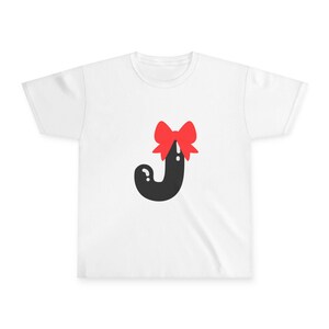 Op de afbeelding: Wit t-shirt met een zwarte letter "J" en een rode strik. De afbeelding staat in het midden van de voorkant van het shirt. Het t-shirt heeft korte mouwen en een ronde hals.