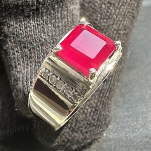 Pode incluir: Um anel de prata com uma grande pedra vermelha vibrante de corte quadrado. A banda do anel é adornada com uma fileira de pequenos diamantes brilhantes. O design é clássico, realçando o brilho da gema.