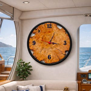 May include: A large, round wall clock with a natural wood grain face and black numerals. The clock has a black frame and red second hand. The clock is mounted on a white wall inside a boat, with a view of the ocean.