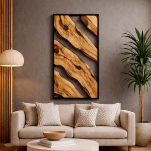 May include: A framed wooden wall art piece featuring natural wood grain patterns in shades of brown and tan. The artwork is encased in a black frame and hangs on a gray wall. The room also includes a beige sofa, pillows, and a potted plant.