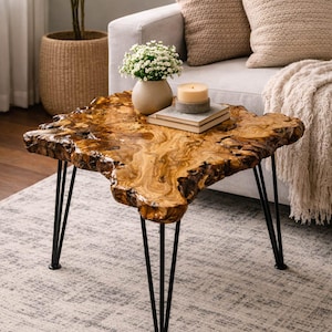Pode incluir: Mesa de centro com borda natural e acabamento em grão de madeira. A mesa tem uma forma orgânica única e é suportada por pernas de metal pretas. Um vaso de flores brancas, uma vela e livros estão sobre a mesa.