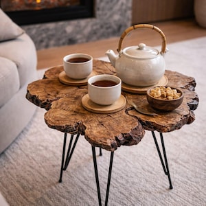 Op de afbeelding: Een rustieke salontafel van hout met zwarte metalen poten. Twee witte theekopjes met thee staan op houten onderzetters, naast een witte theepot met een bamboe handvat. Er staat ook een kleine kom met bruine suikerklontjes.