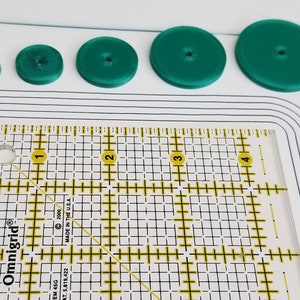 Peut inclure: Cinq cercles en plastique vert de tailles diff&eacute;rentes, avec une r&egrave;gle dans le coin inf&eacute;rieur gauche. La r&egrave;gle est gradu&eacute;e en pouces et comporte une grille.