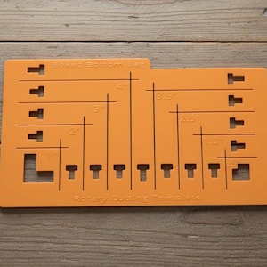 May include: A rectangular orange plastic sewing template for boxed bottoms with black printed markings and cutouts. The tool features various measurements in inches, including 1.5, 2, 2.5, 3, 3.5, and 4 inches, with the text Boxed Bottom Bag and Rotary Cutting Template.