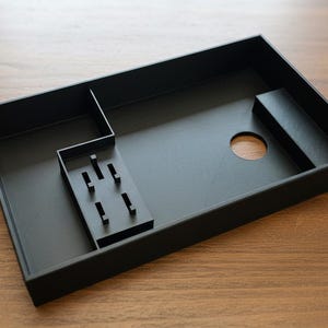 May include: A rectangular black organizer tray featuring multiple internal compartments, a circular cutout, and a small raised section with six vertical prongs for holding items. The tray has a matte finish and a structured, open-top design.
