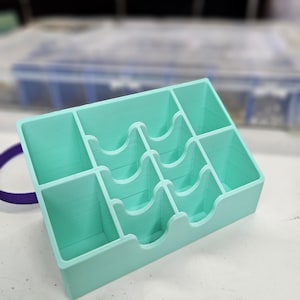 May include: A rectangular, light teal-colored organizer with multiple compartments. The organizer has a series of curved dividers and is designed to hold small items. The item is made of a solid material and sits on a white surface.