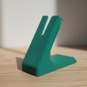 May include: A solid teal green plastic stand featuring an angled support structure with a vertical slot cut into the top center. The object has a flat base and a textured surface finish, designed with a geometric shape for holding items securely.