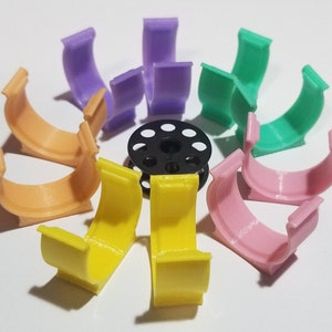 May include: Assortment of colorful plastic thread spool holders in various colors, including orange, yellow, pink, green, and purple. The holders are designed to hold spools of thread for sewing or crafting.