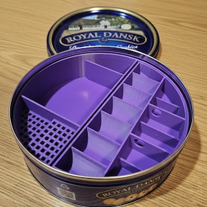 May include: A purple organizer insert inside a Royal Dansk cookie tin. The insert has multiple compartments of varying sizes. The tin's lid is open, revealing a blue and gold design. The tin is on a wooden surface.
