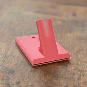 May include: A coral-colored plastic stand featuring a flat rectangular base with a small circular hole and an upright, V-shaped support arm extending from the center. The object has a matte finish and a uniform, solid color throughout its structure.