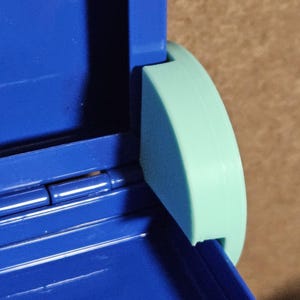 May include: Close-up of a blue plastic container with a hinged lid and a rounded, light green handle. The hinge is silver. The container is on a brown surface.