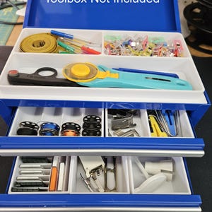 May include: A blue metal toolbox with two drawers. The top drawer is open and contains various sewing supplies, including a tape measure, scissors, and a rotary cutter. The bottom drawer is closed. The text "Toolbox Not Included" is printed on the top of the toolbox. The text "Sample Items Only - Not Included" is printed on the bottom of the toolbox.
