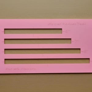 Op de afbeelding: Een rechthoekig roze plastic ritszakgereedschap voor #5 ritsen met vier horizontale uitsparingen gelabeld met 15,24 cm, 17,78 cm, 20,32 cm en 22,86 cm om te helpen bij naai- en knutselprojecten.