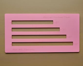 May include: A rectangular pink plastic Zipper Pocket Tool for #5 zippers featuring four horizontal cutouts labeled with measurements of 6 inches, 7 inches, 8 inches, and 9 inches to assist with sewing and crafting projects.