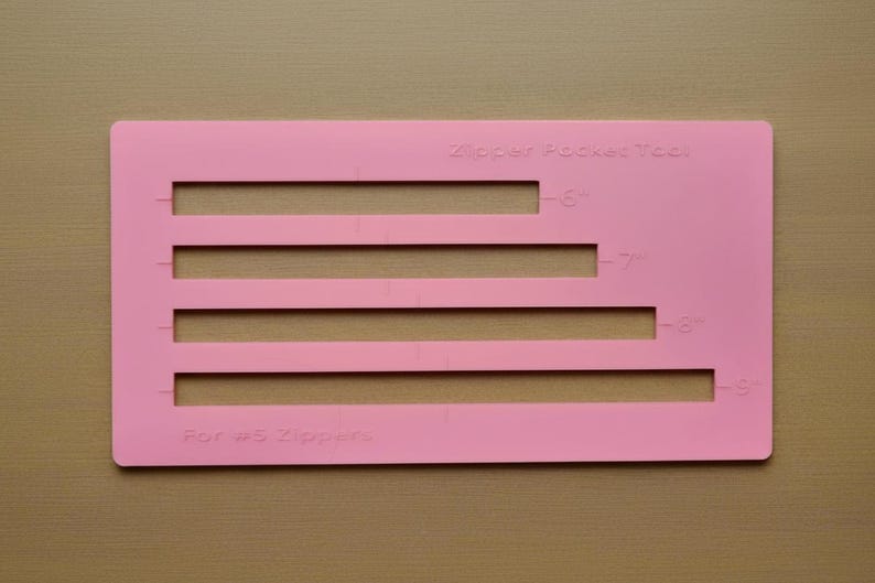 Op de afbeelding: Een rechthoekig roze plastic ritszakgereedschap voor #5 ritsen met vier horizontale uitsparingen gelabeld met 15,24 cm, 17,78 cm, 20,32 cm en 22,86 cm om te helpen bij naai- en knutselprojecten.