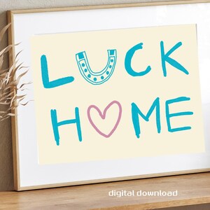 Puede incluir: Impresión artística enmarcada con las palabras "LUCK HOME" en turquesa y rosa. La palabra "LUCK" tiene una herradura encima. La impresión está sobre un fondo crema y se muestra en una estantería de madera con objetos decorativos.