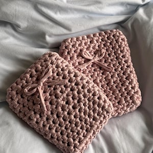 Könnte beinhalten: Zwei handgefertigte, quadratische, blushfarbene Häkeltaschen mit einem strukturierten Stichmuster. Jede Tasche hat ein passendes Band, das zu einer Schleife gebunden ist. Die Taschen werden auf einem hellblauen Stoffhintergrund präsentiert.