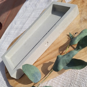 Könnte beinhalten: Ein rechteckiger, hellgrauer Beton-Pflanzkübel mit einem herzförmigen Detail. Der Pflanzkübel liegt auf einem Holzbrett, mit einem Zweig Eukalyptusblättern. Der Pflanzkübel ist etwa 30 cm lang.