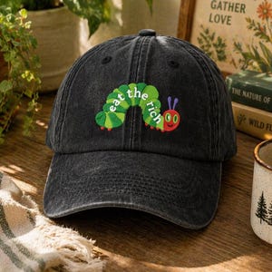 Può includere: Cappellino da baseball nero con la scritta ricamata "eat the rich" in verde e bianco, con un bruco dei cartoni animati. Il cappellino ha una visiera curva e un bottone sulla parte superiore. Il design ricorda un libro per bambini.