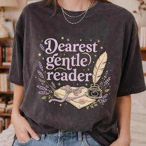 May include: Dark gray t-shirt with the text "Dearest gentle reader" in a whimsical font, accompanied by illustrations of a feather quill, inkwell, and a stack of books tied with a bow. Lavender sprigs and stars complete the design.