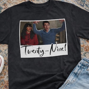 Può includere: T-shirt nero con grafica fotografica di due persone e la scritta "Twenty-Nine!". La maglietta è abbinata a jeans blu e scarpe da ginnastica bianche. È visibile anche una tazza di caffè.