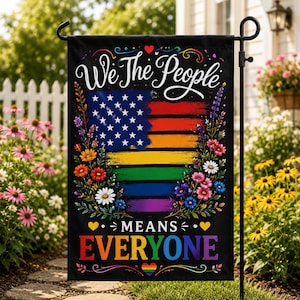 May include: A black garden flag featuring the white text We The People at the top and MEANS EVERYONE at the bottom. The center displays a stylized American flag with rainbow-colored stripes and white stars, framed by colorful floral illustrations.