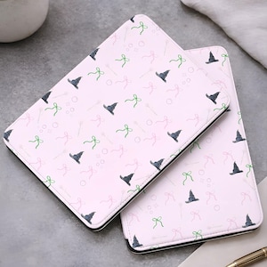 Może przedstawiać: Dwa różowe etui na tablet z powtarzającym się wzorem szarych kapeluszy czarownic, zielonych kokardek i różowych kokardek. Etui mają czarną obwódkę. Tło jest neutralnie szare.