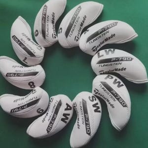 以下が含まれることがあります： 緑色の表面に円形に配置された白いゴルフクラブヘッドカバーのコレクション。各カバーには「P.790 TUNGSTEN TaylorMade」の文字と、「LW」、「PW」、「SW」などのクラブ指定または番号が記載されています。