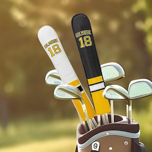May include: Golf club head covers in a golf bag. One cover is white with yellow and black accents, the other is black with yellow accents. Both covers have the name "Gilmore" and the number "18" printed on them.