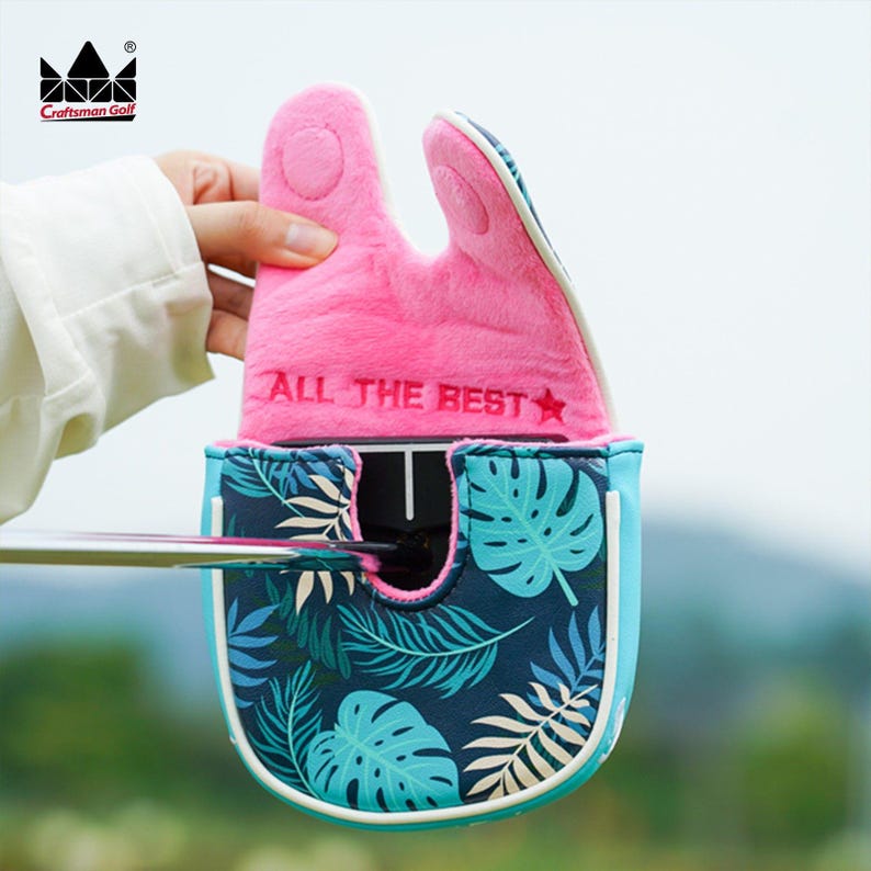 K&ouml;nnte beinhalten: Eine Golf-Putter-Schl&auml;gerhaube mit einem rosa, pl&uuml;schigen Oberteil und einem blau-gr&uuml;nen tropischen Bl&auml;ttermuster. Die H&uuml;lle hat den Text "ALL THE BEST" und einen Stern. Das Craftsman Golf-Logo befindet sich oben links.