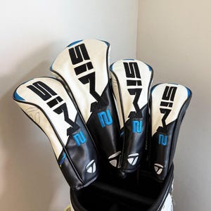 May include: A set of four golf club head covers. The covers are white with black and blue detailing. The text "SIM" and the number "2" are printed on the covers. The covers are in a black golf bag.