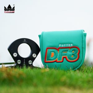 K&ouml;nnte beinhalten: Ein schwarzer Golf-Putter mit kreisf&ouml;rmigem Design und silbernen Schrauben, neben einer tealfarbenen Putterhaube mit dem Text "PUTTER DF3" in Orange und Rot. Das Craftsman Golf Logo befindet sich oben links.