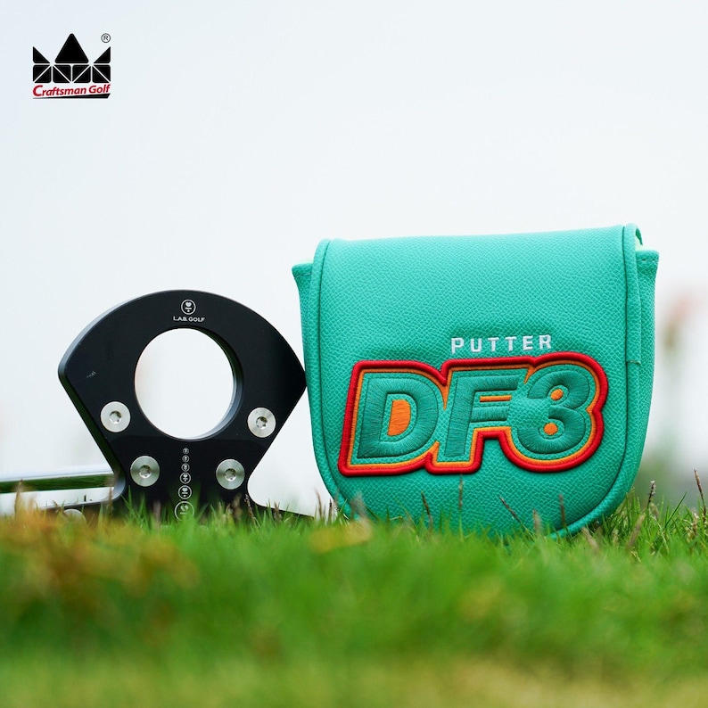 K&ouml;nnte beinhalten: Ein schwarzer Golf-Putter mit kreisf&ouml;rmigem Design und silbernen Schrauben, neben einer tealfarbenen Putterhaube mit dem Text "PUTTER DF3" in Orange und Rot. Das Craftsman Golf Logo befindet sich oben links.