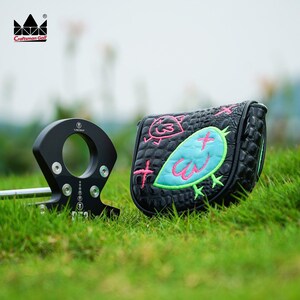 K&ouml;nnte beinhalten: Schwarzer Golfschl&auml;ger und Schl&auml;gerhaube auf gr&uuml;nem Gras. Die Schl&auml;gerhaube ist schwarz mit einem t&uuml;rkis-rosa Vogeldesign und einer Krokodilhautstruktur. Das Craftsman Golf-Logo befindet sich oben links.