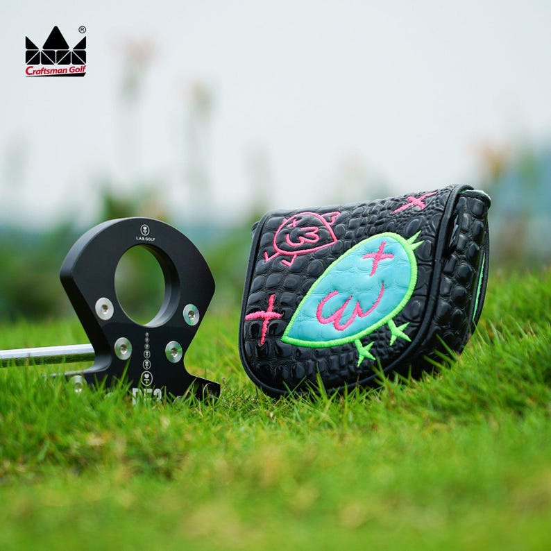 K&ouml;nnte beinhalten: Schwarzer Golfschl&auml;ger und Schl&auml;gerhaube auf gr&uuml;nem Gras. Die Schl&auml;gerhaube ist schwarz mit einem t&uuml;rkis-rosa Vogeldesign und einer Krokodilhautstruktur. Das Craftsman Golf-Logo befindet sich oben links.