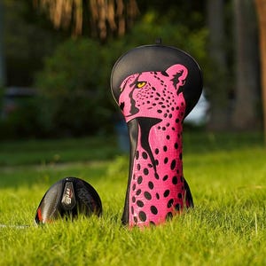 May include: A golf club head cover with a pink leopard print design and a black top. A black and red golf club is in the grass next to the cover. The cover is standing upright in the green grass.