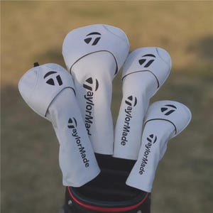 Puede incluir: Un juego de cuatro fundas blancas para la cabeza de palos de golf con el logotipo TaylorMade en negro. Las fundas están hechas de un material suave y están diseñadas para proteger los palos de golf. El fondo es verde borroso.