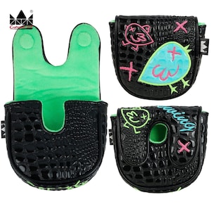 K&ouml;nnte beinhalten: Schwarze Golf-Putter-H&uuml;llen mit Krokodiltextur und gr&uuml;nem Innenraum. Eine H&uuml;lle zeigt ein Vogeldesign mit rosa und blauen Akzenten. Eine andere H&uuml;lle hat das Wort "swinging" in Blau. Die H&uuml;llen haben einen Magnetverschluss.
