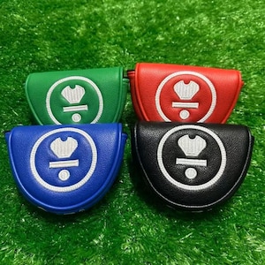 May include: Four golf club head covers in green, red, blue, and black. Each cover has a white embroidered emblem within a white circle. The covers are arranged on a green surface, possibly artificial turf.