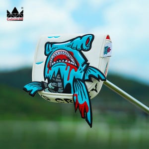 K&ouml;nnte beinhalten: Eine wei&szlig;e Golfschl&auml;gerhaube mit einem Cartoon-Hai-Design in Blau und Rot. Der Hai hat ein offenes Maul mit sichtbaren Z&auml;hnen und Blut, das heruntertropft. Die H&uuml;lle hat das Logo "Craftsman Golf".