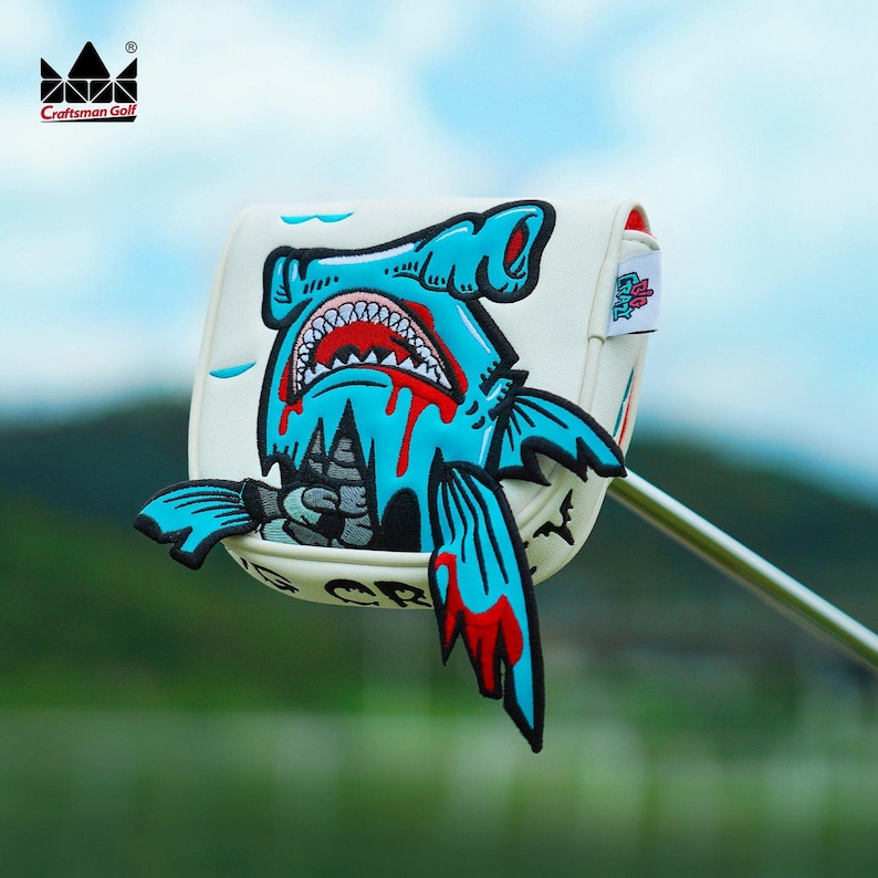 K&ouml;nnte beinhalten: Eine wei&szlig;e Golfschl&auml;gerhaube mit einem Cartoon-Hai-Design in Blau und Rot. Der Hai hat ein offenes Maul mit sichtbaren Z&auml;hnen und Blut, das heruntertropft. Die H&uuml;lle hat das Logo "Craftsman Golf".