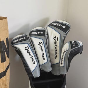 May include: A collection of four golf club head covers. The covers are white, grey, and black with the "SIM" and "TaylorMade" logos. The covers are in a golf bag.