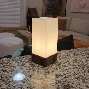 Puede incluir: Una lámpara de mesa rectangular con una luz cálida y brillante. La lámpara tiene una pantalla blanca translúcida y una base de madera marrón oscuro. La lámpara está sobre una encimera de granito.