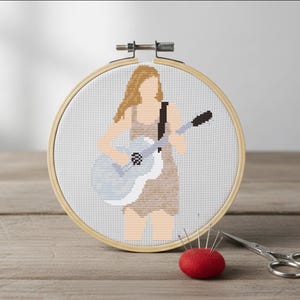 May include: A cross-stitch embroidery in a wooden hoop depicts a person playing a light blue guitar. The figure has blonde hair and is wearing a tan dress. A red pin cushion and small scissors are in the foreground.