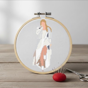 Peut inclure: Un cercle à broder au point de croix représentant une silhouette en tenue blanche et beige avec des cheveux blonds. Un coussin à épingles rouge avec des épingles argentées et de petits ciseaux sont à côté du cercle sur une surface en bois.