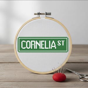 May include: A cross-stitch embroidery hoop displaying "CORNELIA ST" on a green sign. The hoop rests on a wooden surface, accompanied by a red pin cushion and small scissors. The embroidery is in white thread.