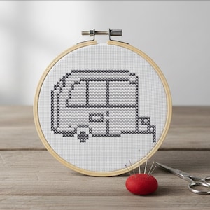 Puede incluir: Un bastidor de bordado de punto de cruz con un diseño de caravana gris sobre tela blanca. Un cojín rojo con agujas y unas pequeñas tijeras plateadas están junto al bastidor. El bastidor de madera se muestra sobre una superficie de madera.