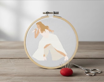 以下が含まれることがあります： 木製の刺繍枠にセットされた、白い衣装を着た人がマイクに向かって歌っているクロスステッチのデザイン。針の付いた赤いピンクッションと小さなハサミが木製の表面にあります。