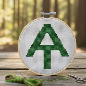 May include: A cross-stitch embroidery hoop featuring a green "AT" design, likely representing the Appalachian Trail. The design is centered on white fabric within a wooden hoop. Green thread and small scissors are also visible.