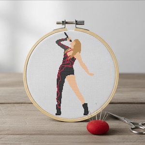 May include: Cross-stitch embroidery in a wooden hoop depicts a person in a black and red outfit holding a microphone. A red pin cushion with needles and a pair of scissors are next to the hoop.